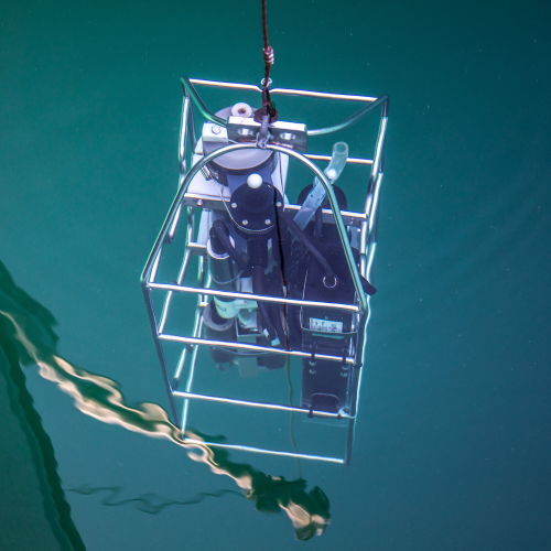 Sonar equipment is lowered into the water. It is surrounded by a metal cage and suspended from a single rope.