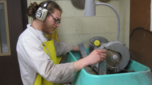 Ludo operates a rotary saw while wearing a yellow apron and ear protectors.