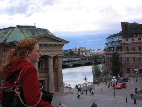 A woman in a red shirt looks off into the distance. Behind her a river runs through the city of Stockholm.