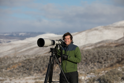Phillipe stands bundled up in front of a camera with a large lens. The hills and shrubs in the background are dusted with snow.