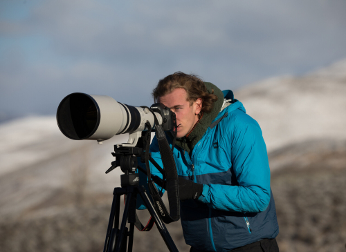 Chris looks through a camera with a large lens. The hills and shrubs in the background are dusted with snow.