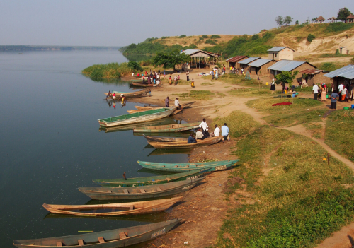 A small village next to a river in Uganda. People go about their lives. Several empty small boats sit on the shore of the river.
