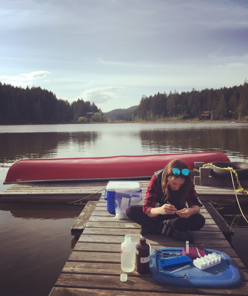 Mackenzie sits cross legged on a dock with an upturned red canoe behind her. She is working with sample collecting equipment.
