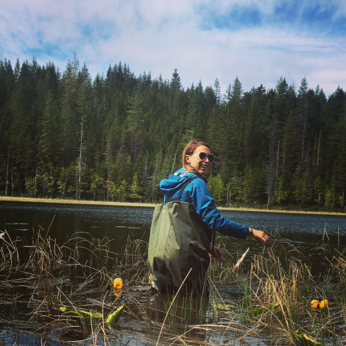 Mackenzie stands in a river dressed in chest waders, a blue jacket and sunglasses. She is only knee deep in water as she smiles back at the camera.
