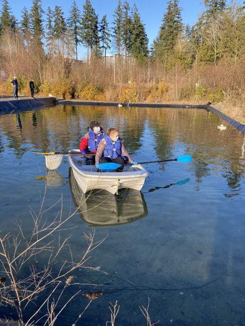 Two researchers wearing life jackets sit in a small boat on an artificial pond lined with black plastic.