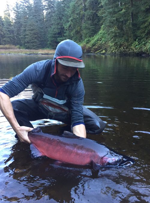 Julian squats in shallow water holding a large red coloured salmon with both hands.