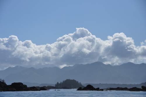 Small islands sit in the foreground. Forested mountains rise in the distance. Fluffy white clouds sit low in the sky with blue sky above them.