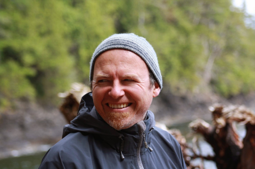 A man smiles as he looks past the camera. He wears a toque and a rain jacket.