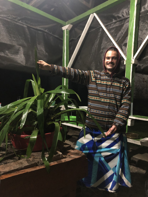 Pierre is under a post-and-beam structure covered by a dark green tarp. It’s night and an overhead lamp illuminates him as he holds up a leaf of a bromeliad that has been moved into a red plastic tub for an experiment.